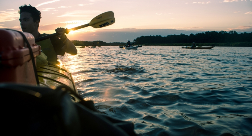 in the foreground, a person paddles a canoe ahead of several others while the sun sets behind them